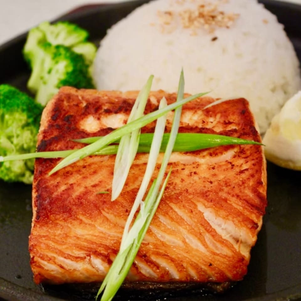 Grilled salmon with green onions, white rice, broccoli, and a lemon wedge on a black plate.