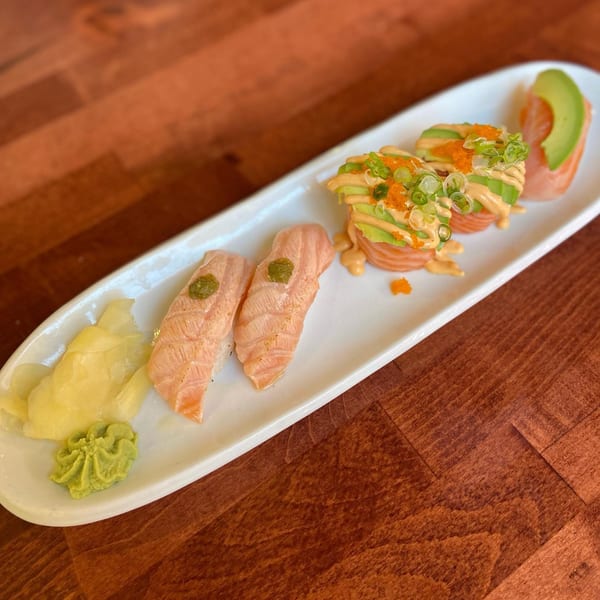 A white platter of salmon nigiri, salmon and avocado rolls, ginger, and wasabi on a wooden table.