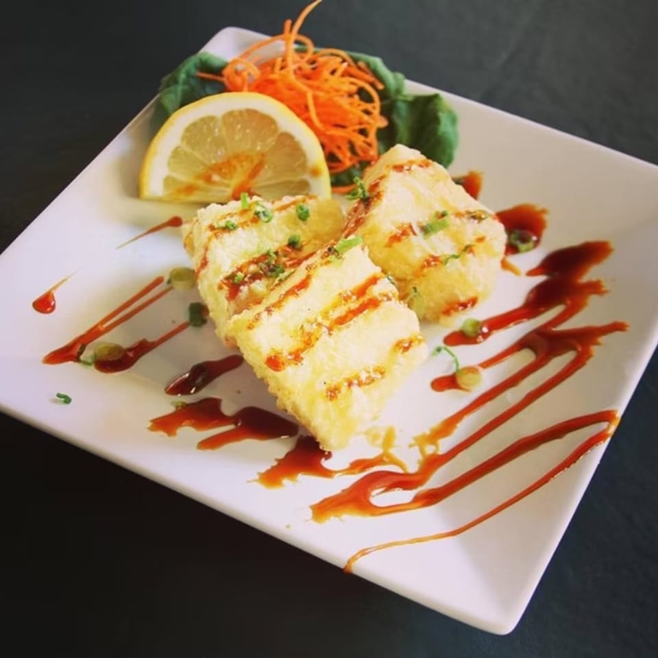 Crispy fried tofu drizzled with sauce, garnished with lemon, shredded carrots, and greens.