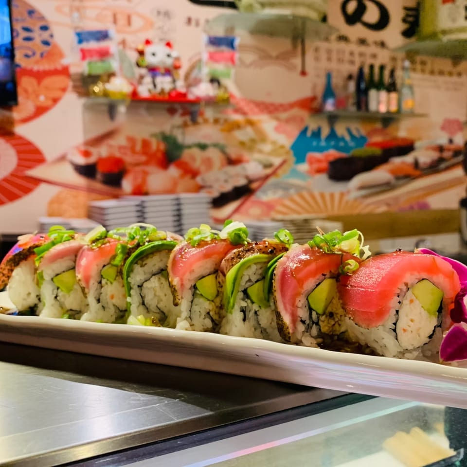 A long plate of sushi rolls with tuna, avocado, and green onions, against a colorful restaurant wall.