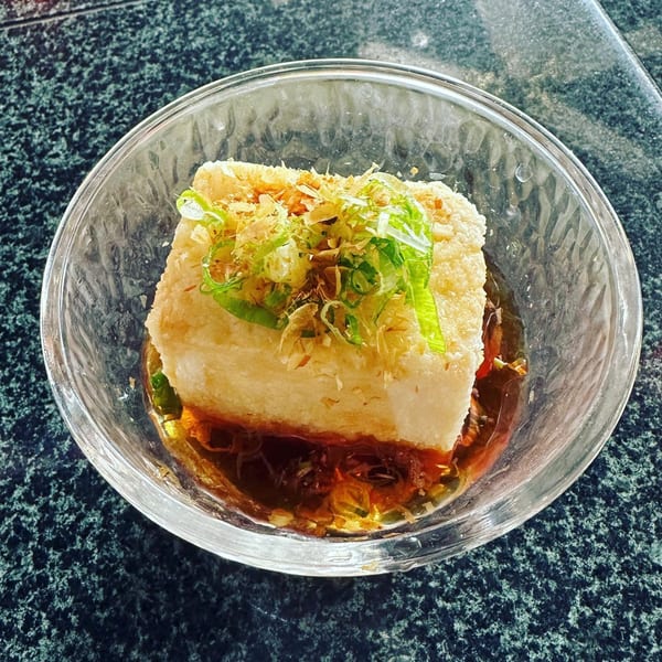 Agedashi tofu in a clear bowl with broth, topped with green onions and bonito flakes.
