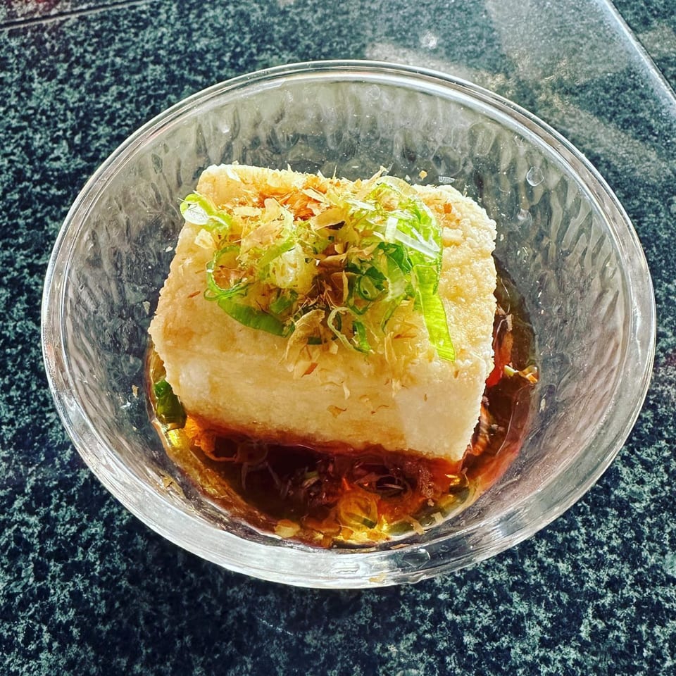 Agedashi tofu in a clear bowl with broth, topped with green onions and bonito flakes.
