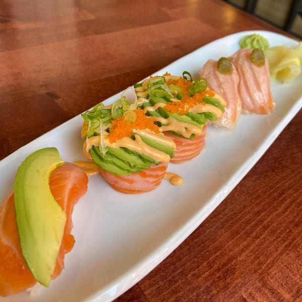 A white rectangular plate of salmon sushi with avocado, roe, and sauce on a wooden table.