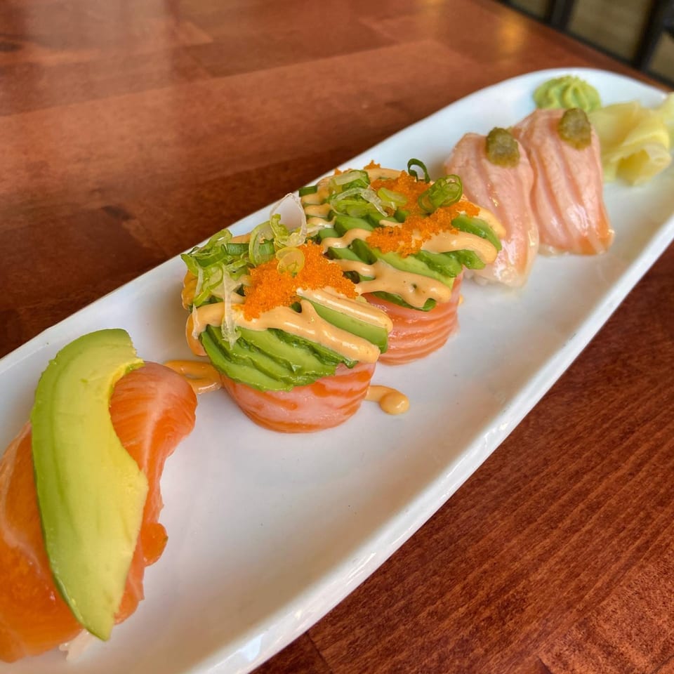 A white rectangular plate of salmon sushi with avocado, roe, and sauce on a wooden table.